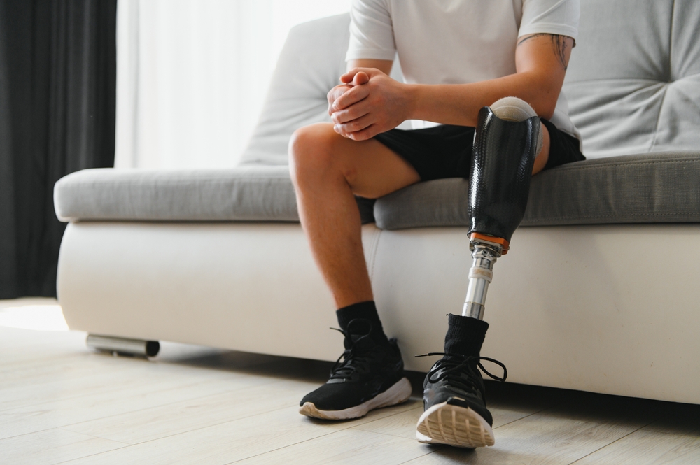 Man amputee with prosthetic leg disability on above knee transfemoral leg prosthesis wearing shoe sitting on sofa, close up. People with amputation disabilities everyday life concept.